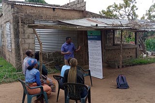 A man stands in front of four people who are sitting in chairs outside a small breezeblock house with a rough corrugated iron verandah. The standing man is speaking and pointing to a display board.