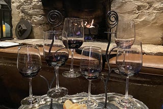 In a wine cellar, 6 wine glasses containing small amounts of red wine displayed on a wire holder, ready to be sampled, with a backdrop of a cozy fireplace.
