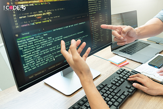 Two developers discussing code on a large monitor in an office setting, with one person pointing at the screen and another gesturing. A keyboard, laptop, smartphone, and notes are visible on the desk. The ICode49 Technolabs logo appears in the top left corner.