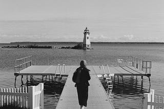 A film photo of a woman in scarf walking towards a pier. We can see a lighthouse in the distance.
