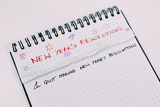 Top spiral bound graph paper with “New Year’s Resolutions” in all capital letters and red ink is written across the top. Small doodles of stars, hearts, and smiley faces in different colored ink surround the title. Below this, many pages have been ripped out, revealing a page with the following written in black ink: “1. Quit Making New Year’s Resolutions”