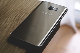 Dark grey Samsung phone laying at the corner of a brown wood desk. Photo by John Tekeridis.
