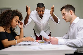 A man angry and gesturing at a table while two people sit and look down at the work that they’ve done, kind of fearful of what that person might say.