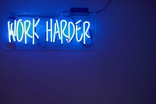 A neon blue sign that says “work harder.”