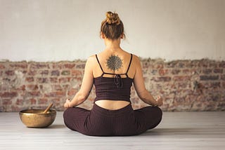 A woman in brown pants and a brown top with a tattoo on her back sits in meditation, looking at a wall. Next to her, a singing bowl sits.