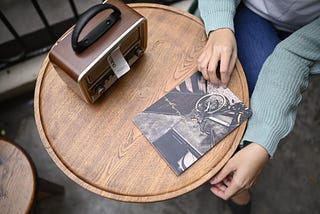 A person sitting at a wooden café table, wearing a green knit sweater, with one hand holding a magazine featuring a bicycle on the cover. A vintage-style portable radio sits on the table.