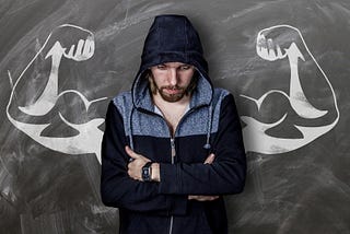 Young white man looking down, and crossing his arms against his chest, wearing a blue and grey hoodie and a serious expression, standing in front of muscular arms drawn on a chalkboard behind him.