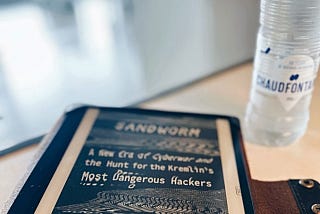An e-reader in a leather case and a bottle of water. The e-reader’s screen shows the book that the reader is currently reading.