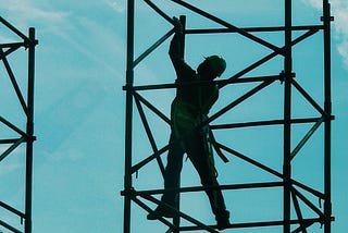 A construction worker stands precariously on scaffolding, suspended in the air, under a blue sky.