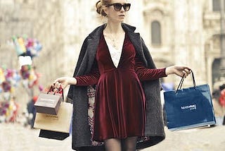 A red-head lady dressed in a burgundy velvet mini-dress wearing sunglasses carries her shopping bags. The background looks like a European street.