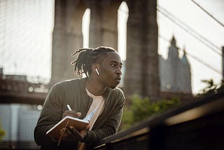 “Positive black man listening to music with wireless earphones and taking notes in diary on street”- Ketut Subiyanto