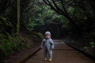 A scared-looking Infant toddler in a grey hooded onesie standing on a wood board path in the middle of a lush green forest.