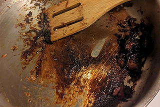 A stainless steel pan with some brown burnt stuff at the bottom and a wooden spatula scraping it up.