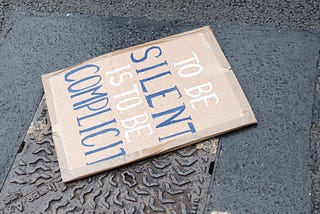 Image of a protest sign on the ground saying “to be silent is to be complicit”