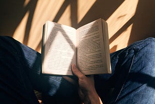 An open book kept between crossed legs of a person who is sitting in sunlight