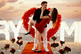 Man holds woman, surrounded by roses and the words “marry me” in lightbulbs.