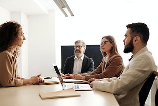 Panel of 3 interviewers, one with a laptop and all in formal dress, sit across from a candidate who is answering a question.