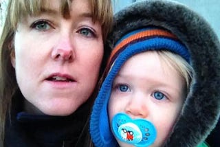 Mother with fringe and blonde baby son with hat and dummy looking up at camera