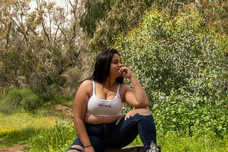 Confident plump girl wearing ripped jeans and crop top sits amongst trees.