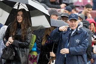 Jerry Jones on the right wearing Dallas Cowboy hat and jacket. Woman in black with black and white umbrella on the left. Random fans in the background.