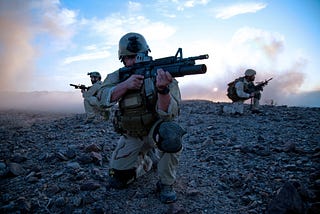 3 Navy SEALs take defensive positions on a beach. The foremost has a M16 with a underbarrel grenade launcher aimed and ready.