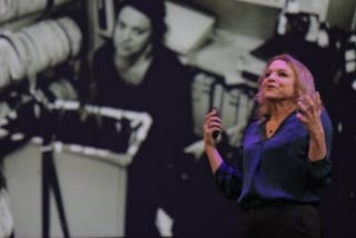 An older white woman with shoulder-length blonde hair stands lectures in front of a black and white image on a movie screen. She wears a blue blouse and slacks.