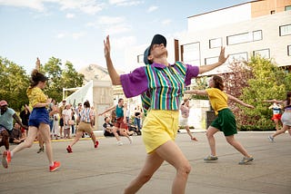 A group of dancers performing outdoors in a plaza. The daner in the foreground wears a striped shirt, baseball cap, and yellow shorts.