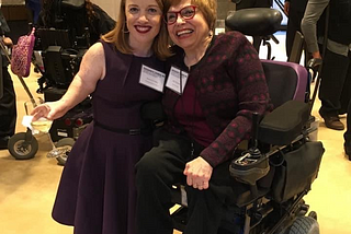 Two women next to each other. One is a little person with shoulder length red hair and is wearing a purple sleeveless dress. The other has short brown hair and glasses is wearing a purple jacket and is seated in a wheelchair. Both are smiling.