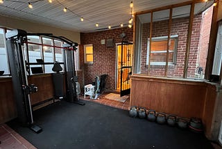 A home gym on a porch, black rubber flooring on most of the ground. A row of kettlebells to the right, a functional trainer with Smith machine on the left.