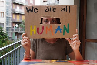 A white, latin girl sitting in an apartment balcolny holding a cardboard sign, “we are all human.” Her face is partially hidden behind the sign with only her eyes visible through a cutout in the cardboard.