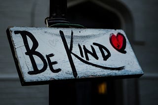 A white rectangular sign on a black post with black letters reading BE KIND with a red heart outlined in black.