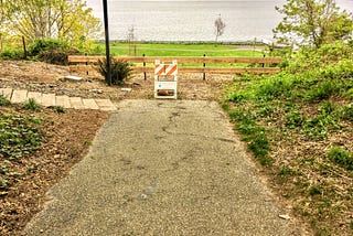 A paved trail dead-ends at a barricade with a new fence and bay in the background