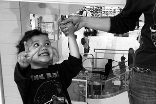 A smily young girl in a shopping mall giving the peace sign while holding her mother’s hand.
