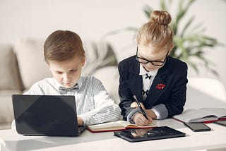 Two children dressed professionally using laptops and tablets indoors.