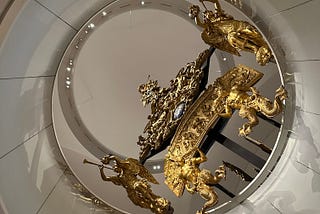 An ornate golden dragon boat sculpture viewed from below, its body and tail curving and twisting around an open circular staircase. The detailed craftsmanship shines under spotlights against the backdrop of the staircase’s simple, modern lines and muted colors.