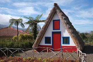A triangular-shaped thatched house with white wall, red door and green shutters; a rustic wooden fence runs in front, and two palm trees.