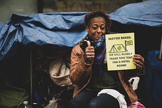 A Black person sits in front of their tent smiling, with their thumb up next to a sign that reads “Mayor Breed we will gladly trade this tent for a hotel room!”