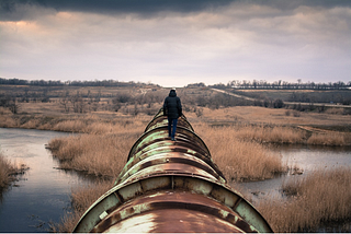 A human figure walks along a rusty pipeline in a bleak, drab, wetland.