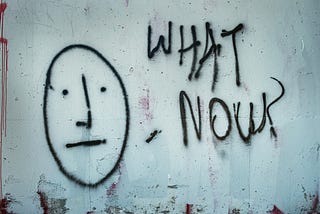 Black graffiti on a white wall — A simple, straight face with the words ‘what now?’