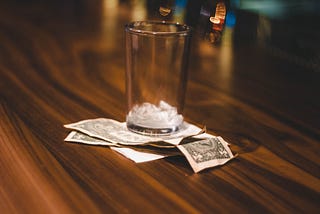 Wood grain bar, with empty 1/4 pint glass, empty of liquid but containing a few ice cubes, sitting atop a stack of two one dollar bills and a paper receipt.