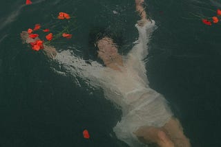 A woman in a white dress floats under water with scattered red petals