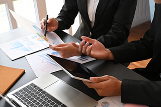 Two people are wearing business suits, and they sit next to each other at an office desk for a meeting. One person holds a piece of paper with charts while the other has a tablet.