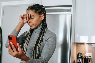 Photograph of a young Black woman with long dreadlocks wearing a gray turtleneck. She stands in her kitchen, holding her phone in one hand, the other is on her forehead. Her eyes are closed. She looks upset.