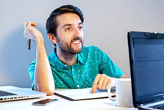 A 20-something man in a teal shirt smiles welcomingly while pointing at a notebook next to his laptop.