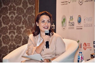 Shymaa enthusiastically holding a microphone, sitting in a chair and speaking.