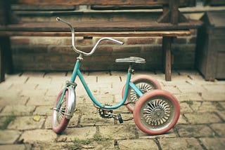 Foto de um velho triciclo azul e com rodas vermelhas. A imagem traz a ideia de uma memória da infância.