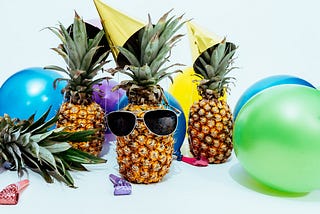 3 pineapples with party accessories surrounded by colourful balloons and noisemakers.