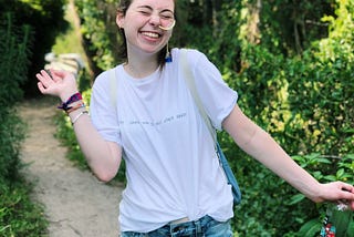 A girl smiling with a white shirt and jean shorts in nature. She has a yellow feeding tube coming out of her nose, running to her backpack, which keeps her nourished.