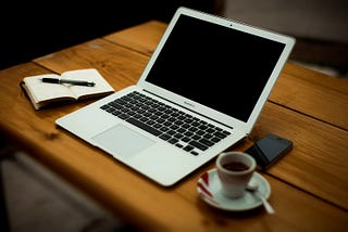 A desk with a laptop, open notebook, and an espresso