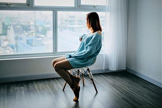 somber girl with brown hair wearing long sleeve blue sweater sitting on chair in empty room looking out the window